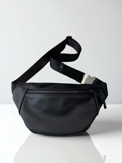 A black pebbled leather fanny pack with an adjustable black webbing strap and a silver metal buckle, displayed on a reflective white surface against a light gray background.