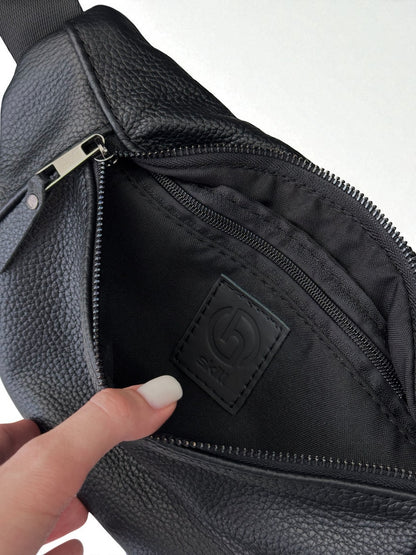 Close-up of a black pebbled leather bag with an open zipper, showing a black interior pocket and a small embossed logo patch. A finger with white nail polish is touching the bag.
