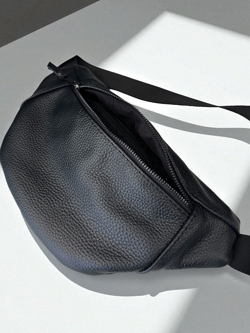 A black pebbled leather belt bag with a zippered top and a black strap, shown on a white surface with dramatic diagonal shadows.