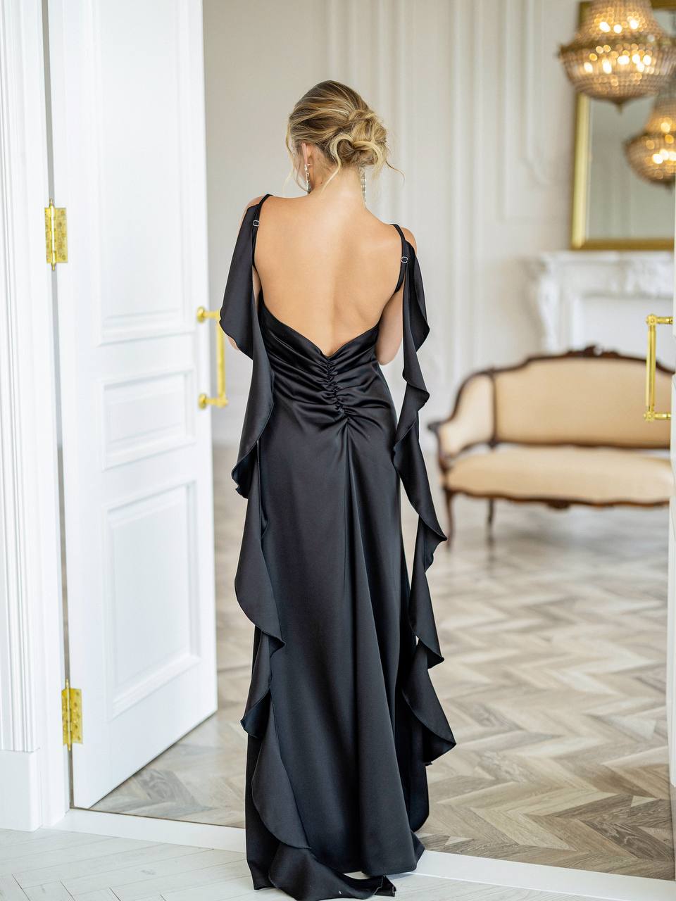 A woman viewed from behind wearing a floor-length black satin gown with a deep open back, ruched center, and dramatic draped side panels, standing in a bright indoor room with a mirror, door, and upholstered bench.