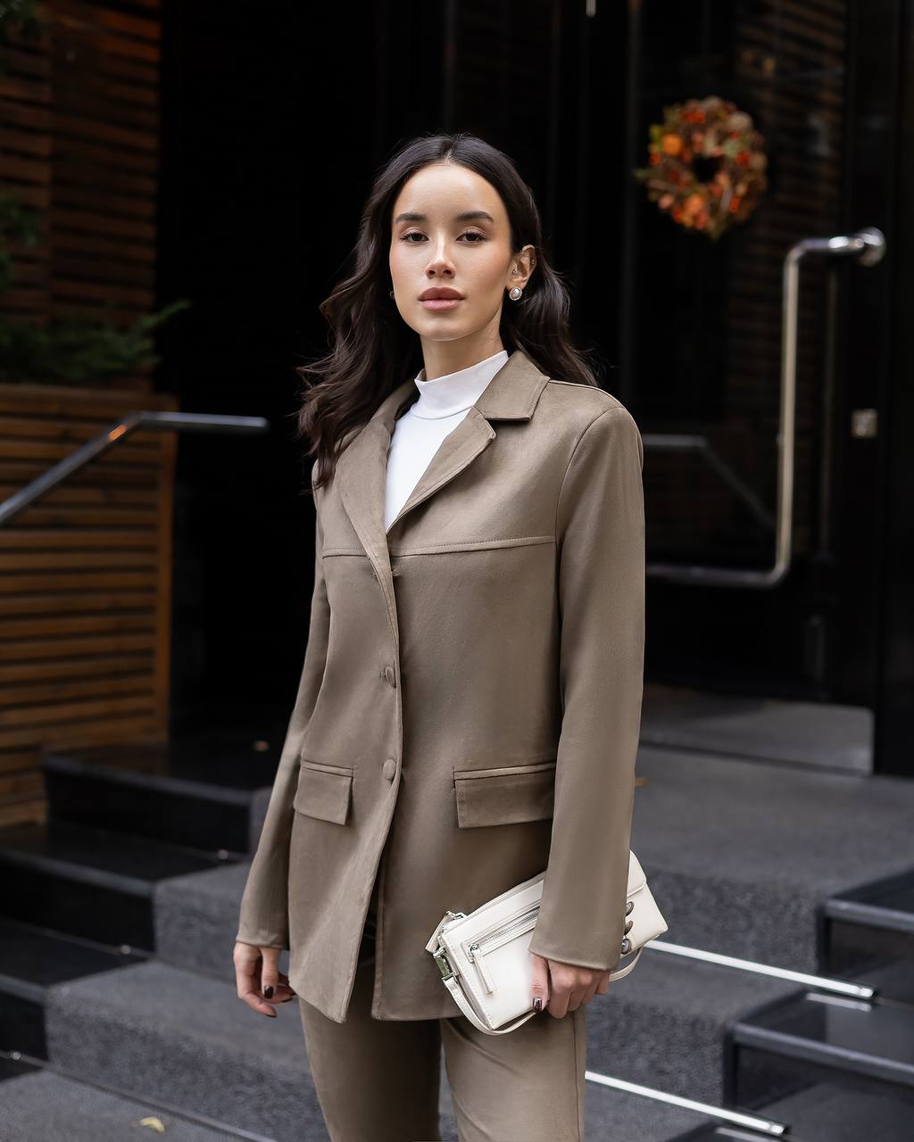 Woman wearing a brown coat and holding a white clutch in an urban setting