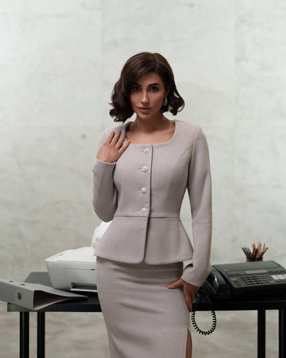 Woman in a light gray suit standing in an office setting with a printer and phone on a desk.