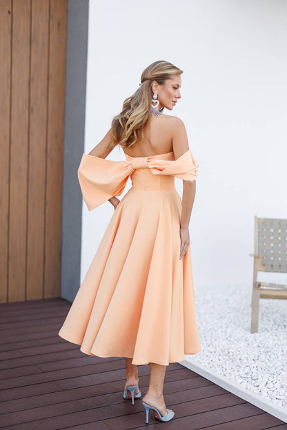 Woman in a peach dress standing on a wooden deck with a white wall and chair in the background.