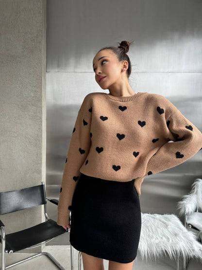 Woman wearing a beige sweater with black heart patterns and a black skirt indoors.