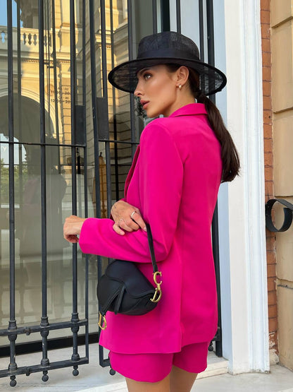 Woman in a bright pink outfit with a black hat and bag, standing in front of a building entrance.