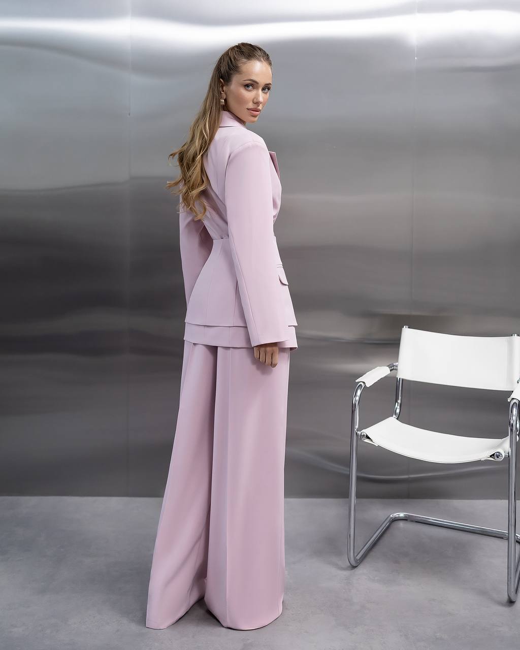 Woman in a pink outfit standing next to a white chair against a reflective silver wall.