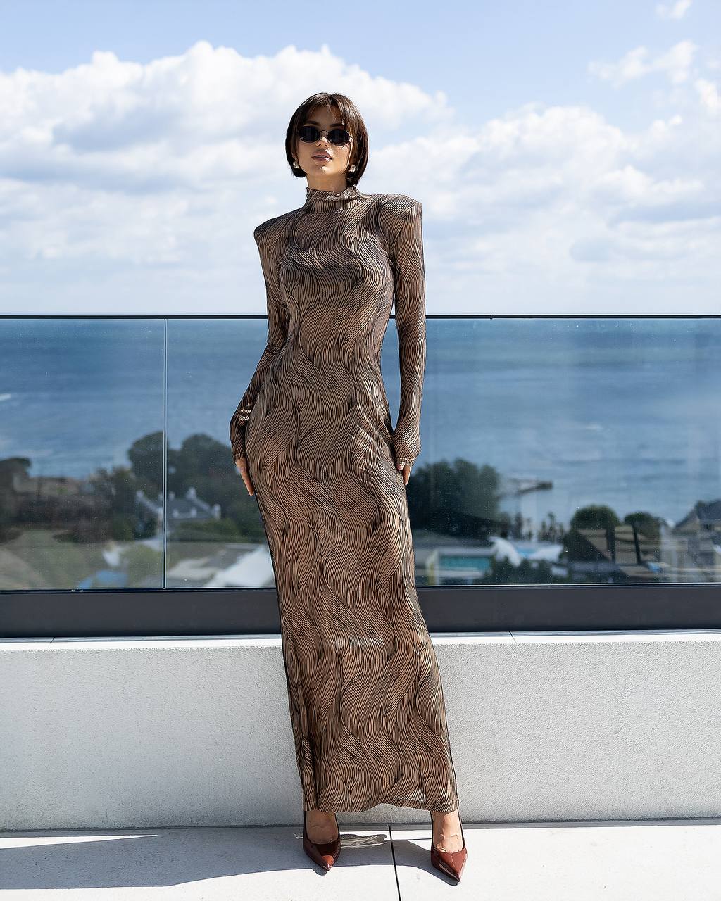 Woman in a long, patterned dress standing on a balcony with a scenic view.