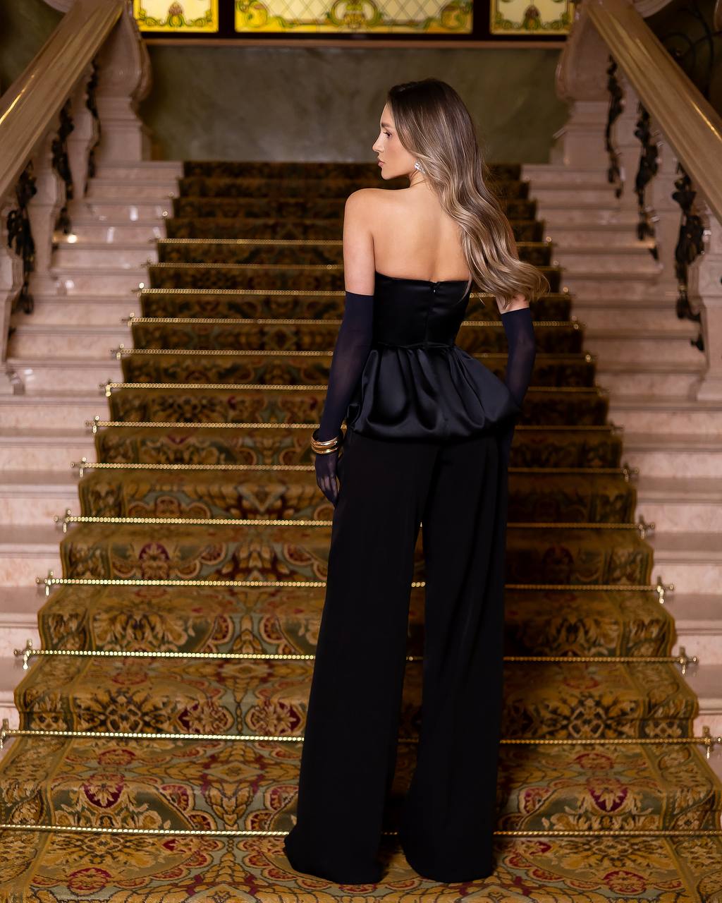 A woman stands on an ornate indoor staircase wearing a black strapless gown with long sheer sleeves and wide-leg pants, photographed from the back.