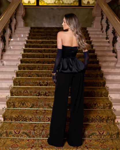 A woman stands on an ornate indoor staircase wearing a black strapless gown with long sheer sleeves and wide-leg pants, photographed from the back.