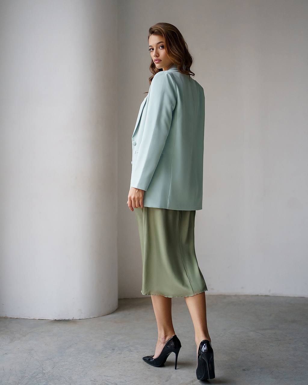 A woman stands in a minimalist studio setting wearing a sage green blazer and a matching sage green midi skirt, paired with black high heels.
