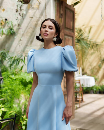 Woman in a light blue dress standing outdoors with greenery and a wooden structure in the background