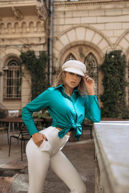 Woman in teal shirt and white pants standing outdoors with a building in the background