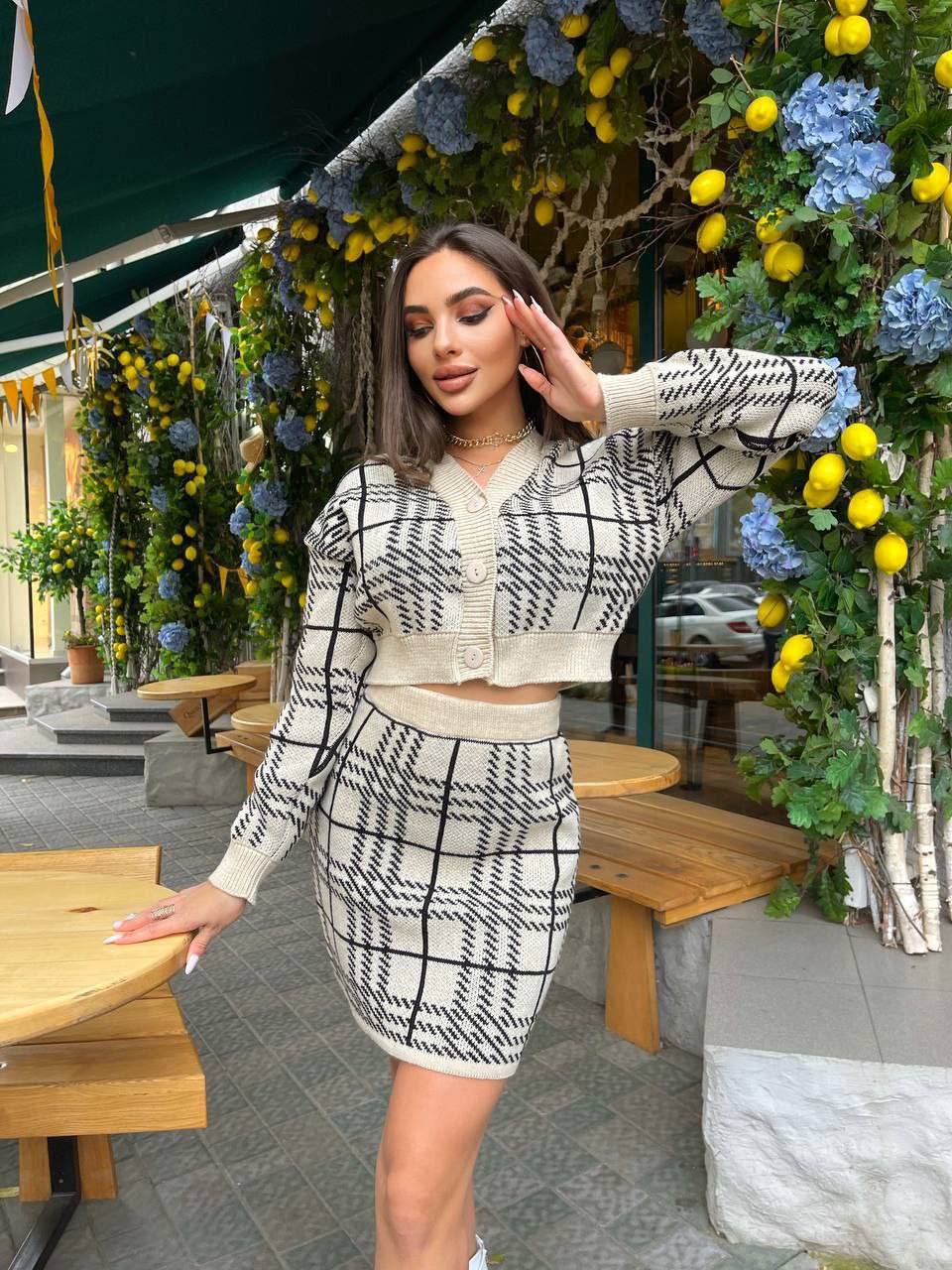 Woman in a checkered outfit standing in an outdoor setting with decorative plants and tables.