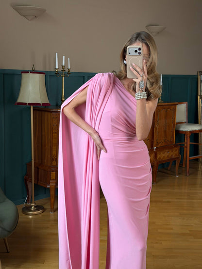 Woman in a pink one-shoulder dress taking a mirror selfie in a room with wooden furniture and a lamp.