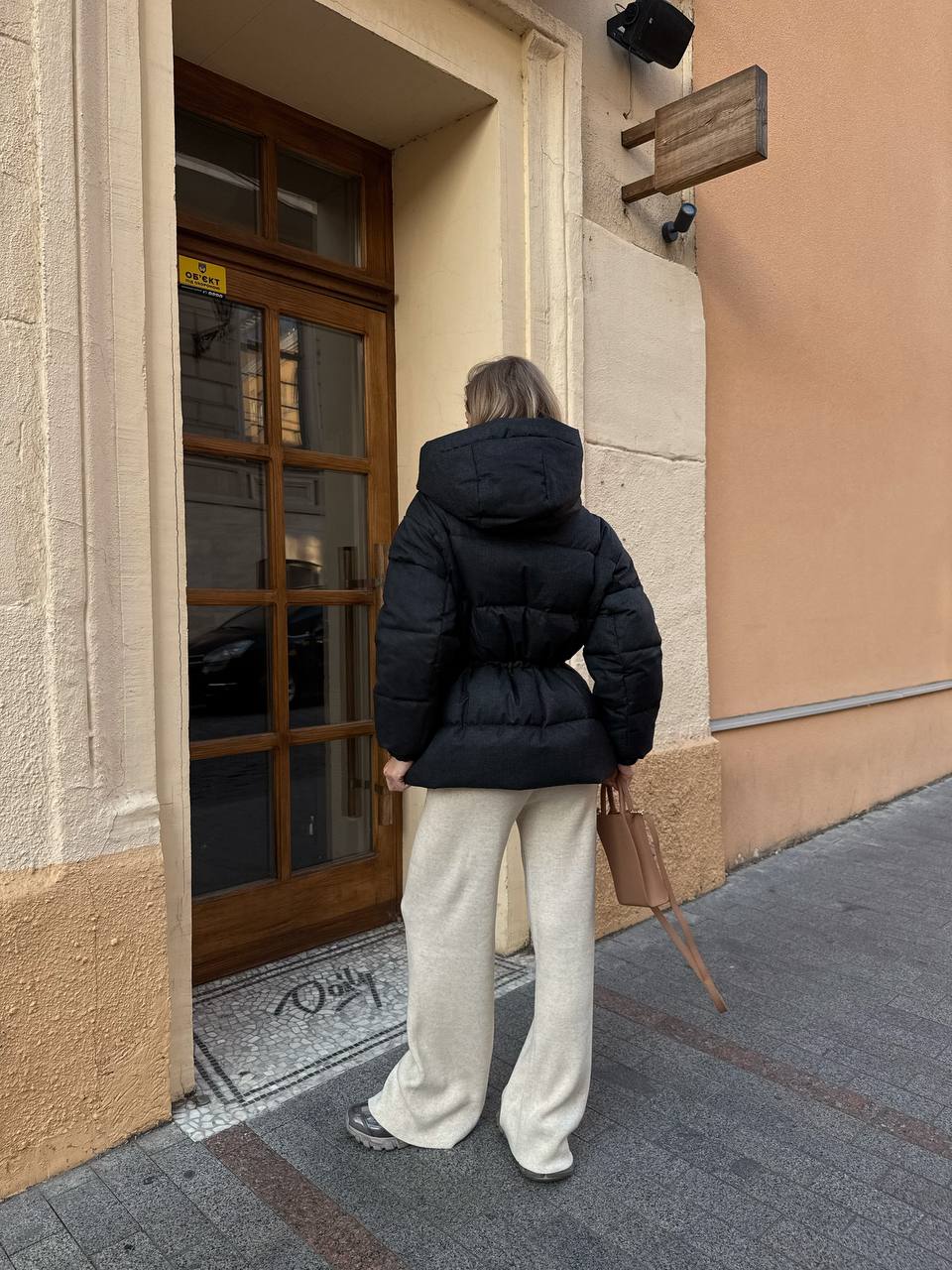 Person wearing a black puffer jacket and white pants standing outside a building.