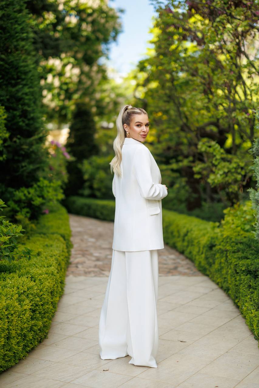 Woman in a white outfit walking through a garden path with greenery on either side.