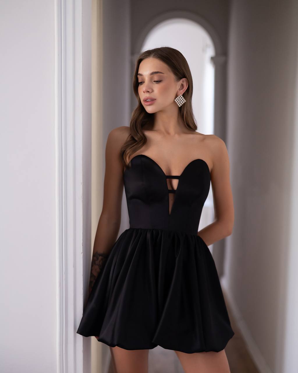A woman modeling a black strapless sweetheart mini dress with a flared skirt, standing in a hallway with white walls and an arched doorway in the background.