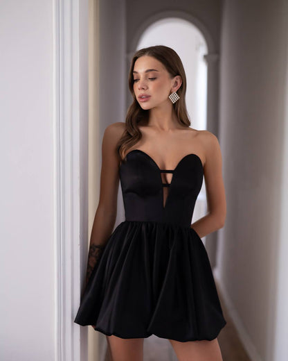 A woman modeling a black strapless sweetheart mini dress with a flared skirt, standing in a hallway with white walls and an arched doorway in the background.