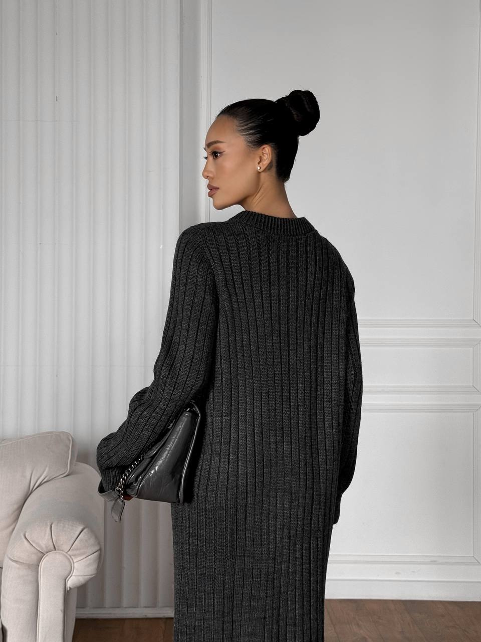 Woman in a black ribbed sweater and skirt standing against a white paneled wall.