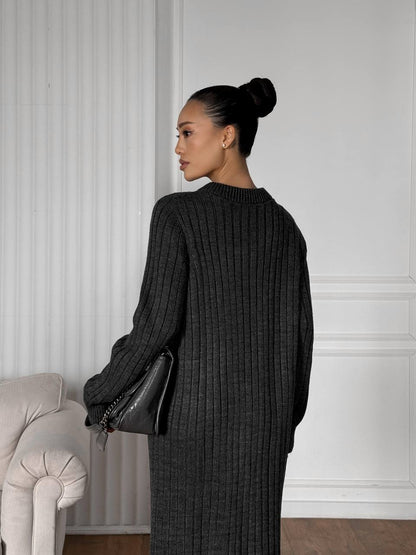 Woman in a black ribbed sweater and skirt standing against a white paneled wall.
