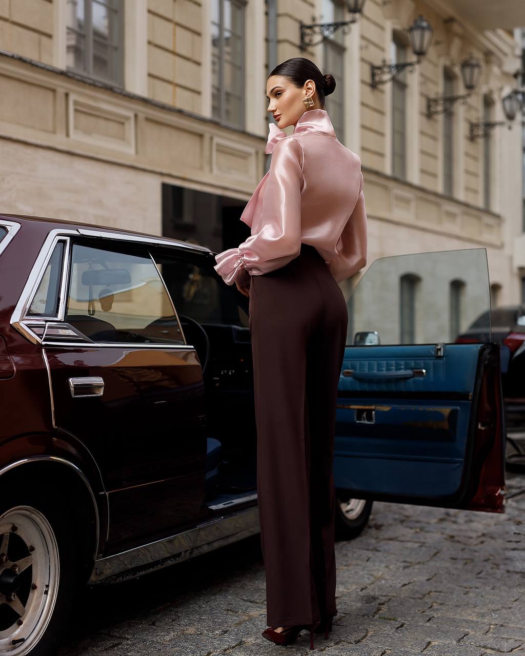 Woman in a pink blouse and brown pants standing next to an open car door on a city street.