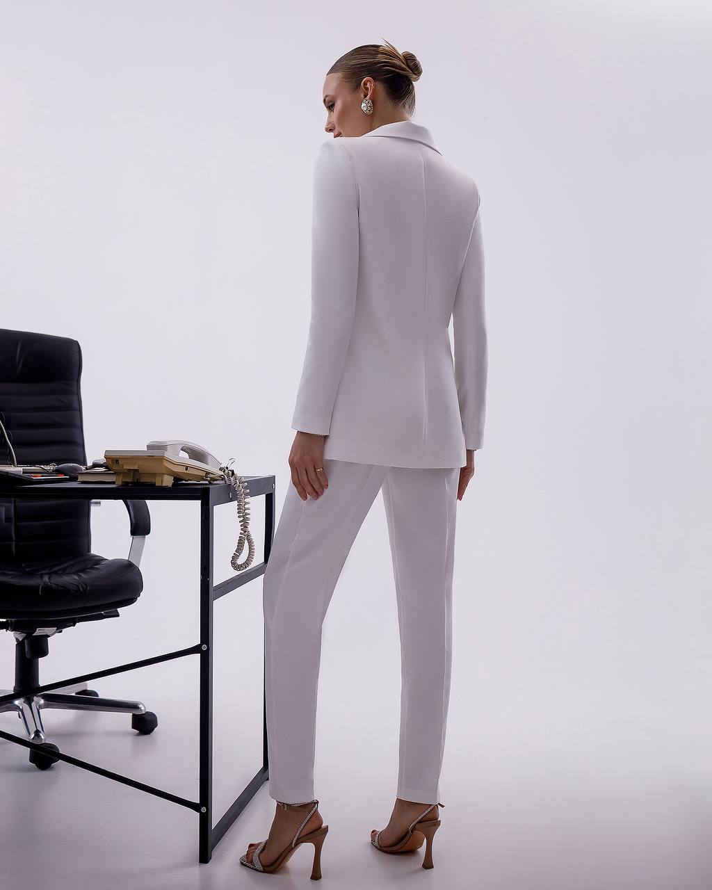 Woman in a white suit standing in an office setting with a desk and chair.