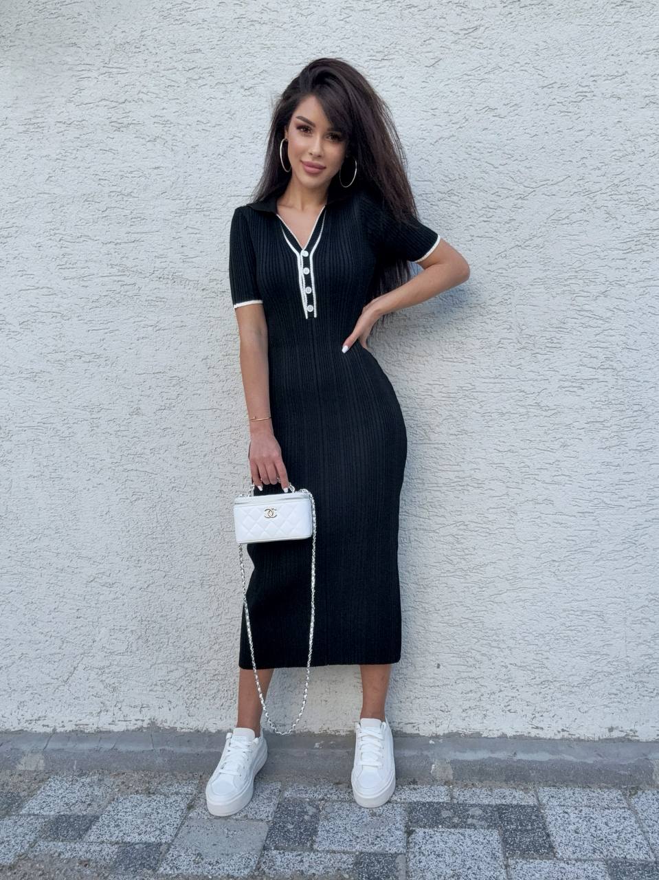 Woman in a black dress with white sneakers and a white handbag against a light gray wall.