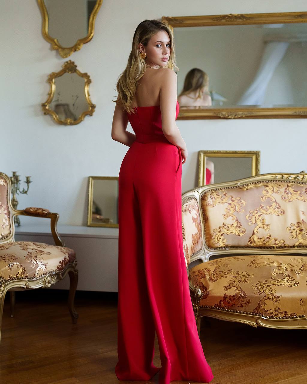 Woman in a red dress standing in an ornate room with mirrors and furniture.