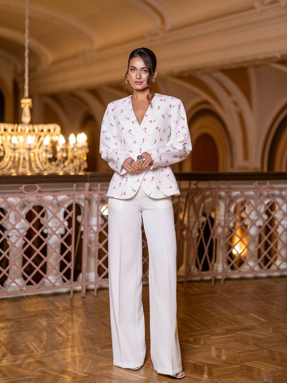 Woman in a white outfit with floral patterns standing in an elegant indoor setting.