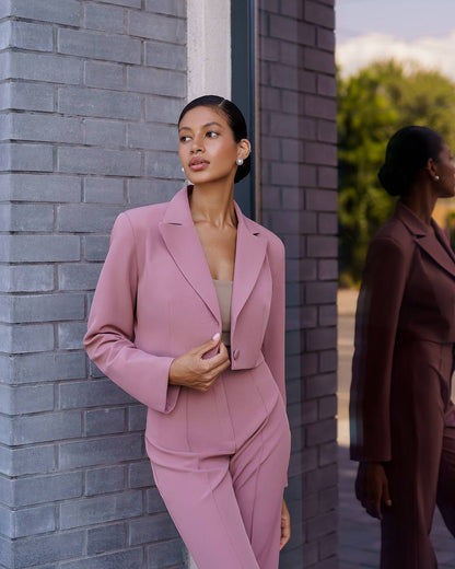 Woman in a pink suit standing against a brick wall with another person in a brown suit in the background.
