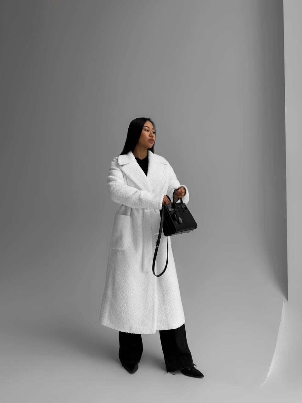 Person wearing a white coat holding a black handbag against a gray background