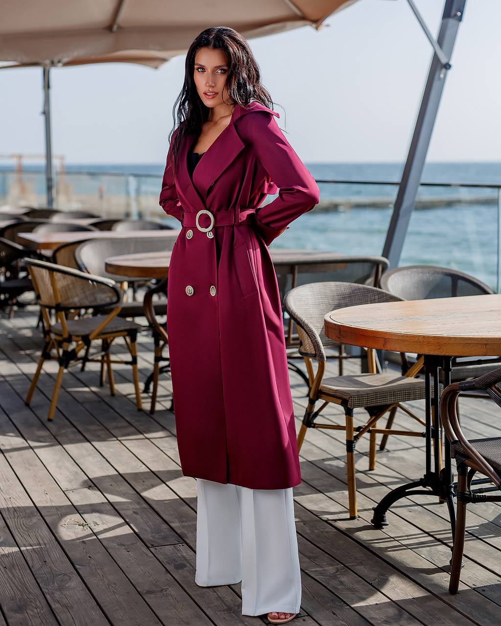 Woman in a red coat standing on a wooden deck with tables and chairs, near water.