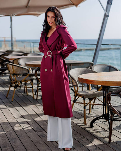 Woman in a red coat standing on a wooden deck with tables and chairs, near water.