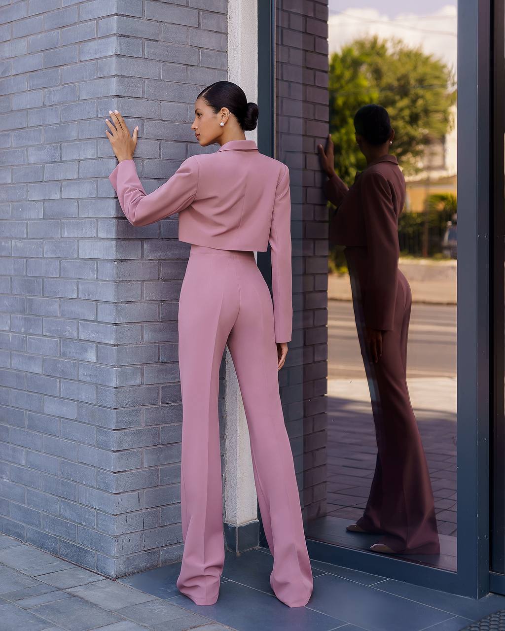 Woman in a pink outfit standing against a brick wall, reflected in a mirror.
