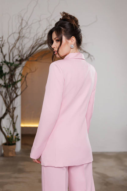 Woman wearing a pink suit standing in a room with a plant and neutral wall.