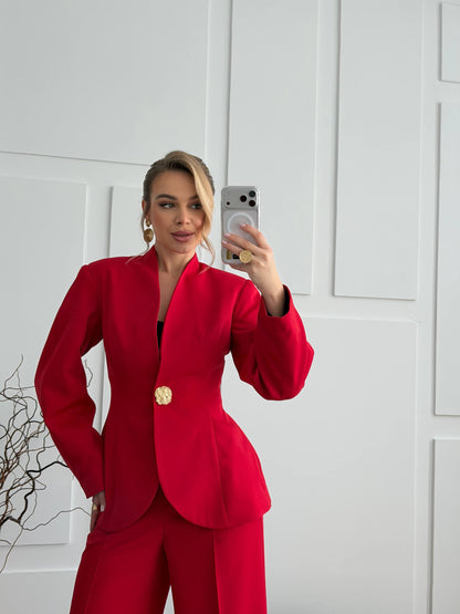 Person wearing a red suit with a gold button, taking a mirror selfie against a white paneled wall.