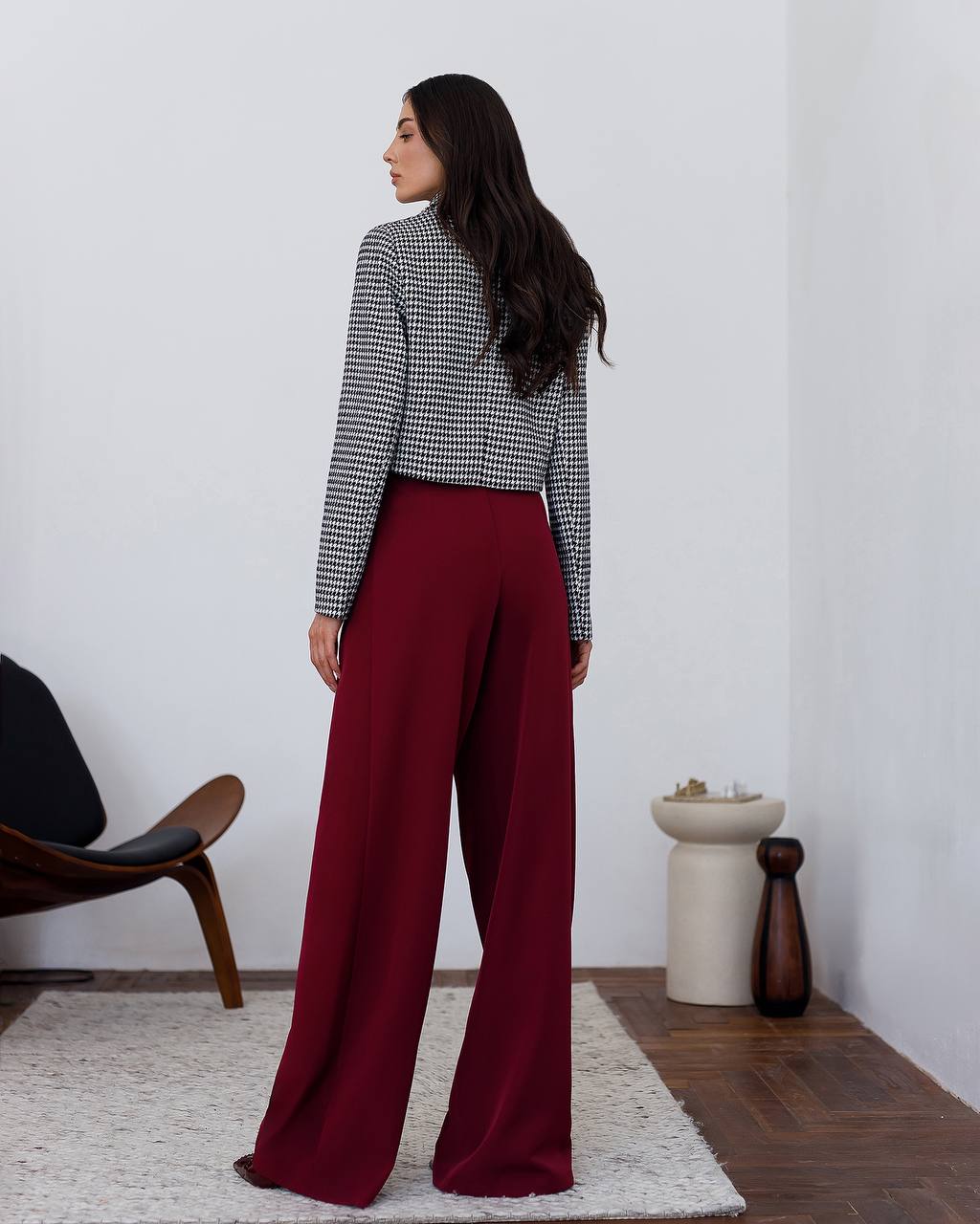 Woman wearing a checkered top and red pants standing in a minimalistic room.