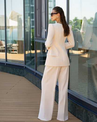A woman stands on a wooden deck beside large glass windows, wearing a light cream or off-white tailored suit with a structured blazer and wide-leg trousers. The photo is taken from behind, showing the back of the outfit and sunglasses.