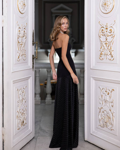 A woman wearing a black strapless maxi dress with a sheer polka-dot overlay stands in an ornate white doorway with gold detailing, looking back over her shoulder.