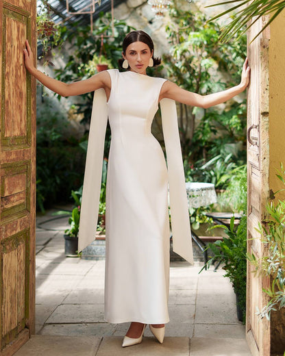 Woman in a white dress standing in a sunlit outdoor setting with plants and a wooden door.