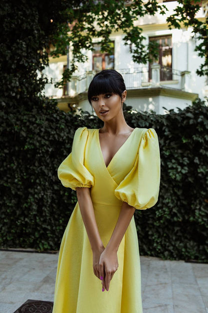 Woman in a yellow dress standing outdoors with greenery and a building in the background