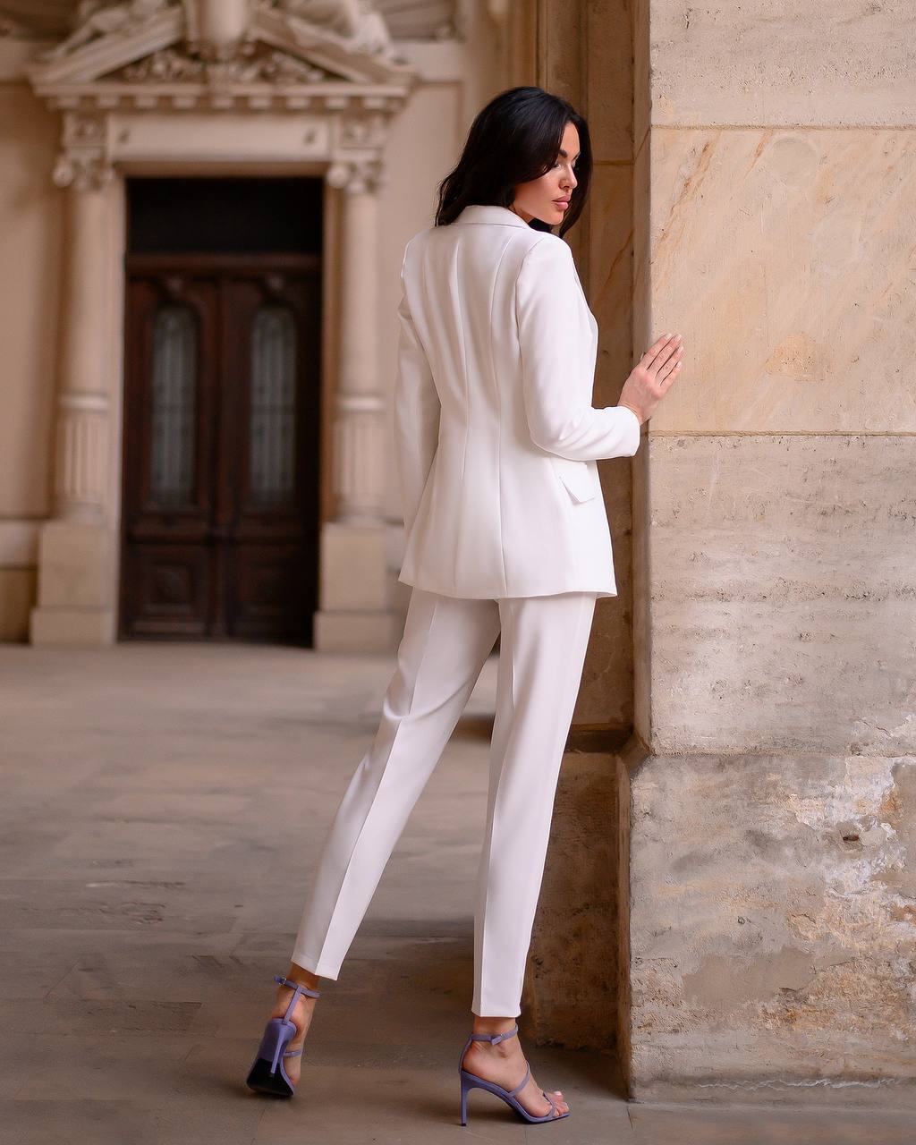 Woman in a white suit standing against a stone wall with classical architecture in the background