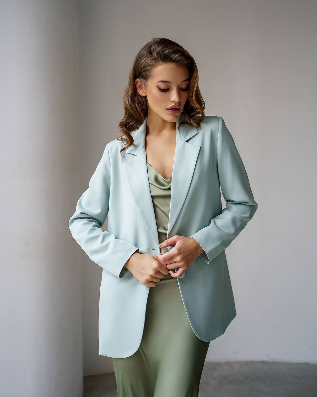 A woman wearing a light sage green blazer over a matching sage green satin top and pants, standing in a minimalist studio setting with a neutral gray background.
