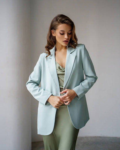 A woman wearing a light sage green blazer over a matching sage green satin top and pants, standing in a minimalist studio setting with a neutral gray background.