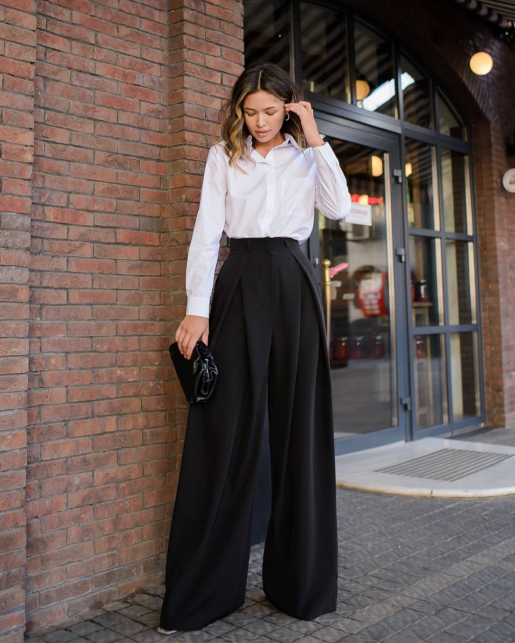 Woman in white shirt and black wide-leg pants standing against a brick wall.