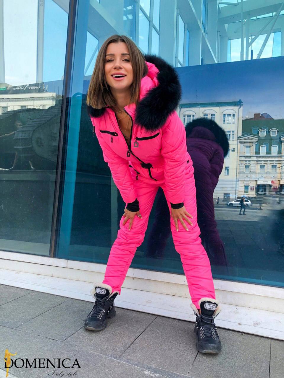 Woman in a bright pink winter outfit with black fur hood standing in front of a glass building.