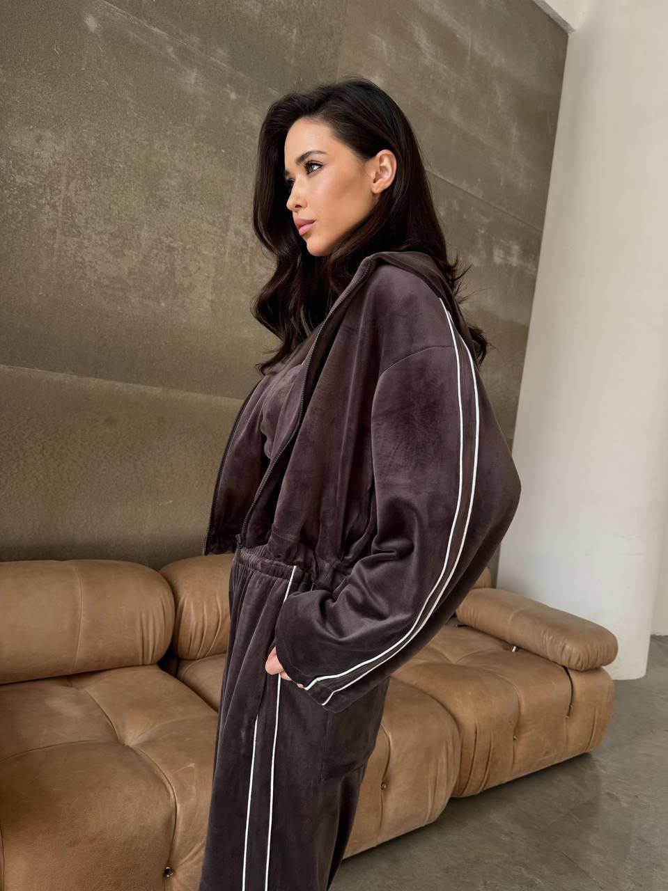 Woman wearing a dark brown tracksuit with white piping standing in a minimalistic room.