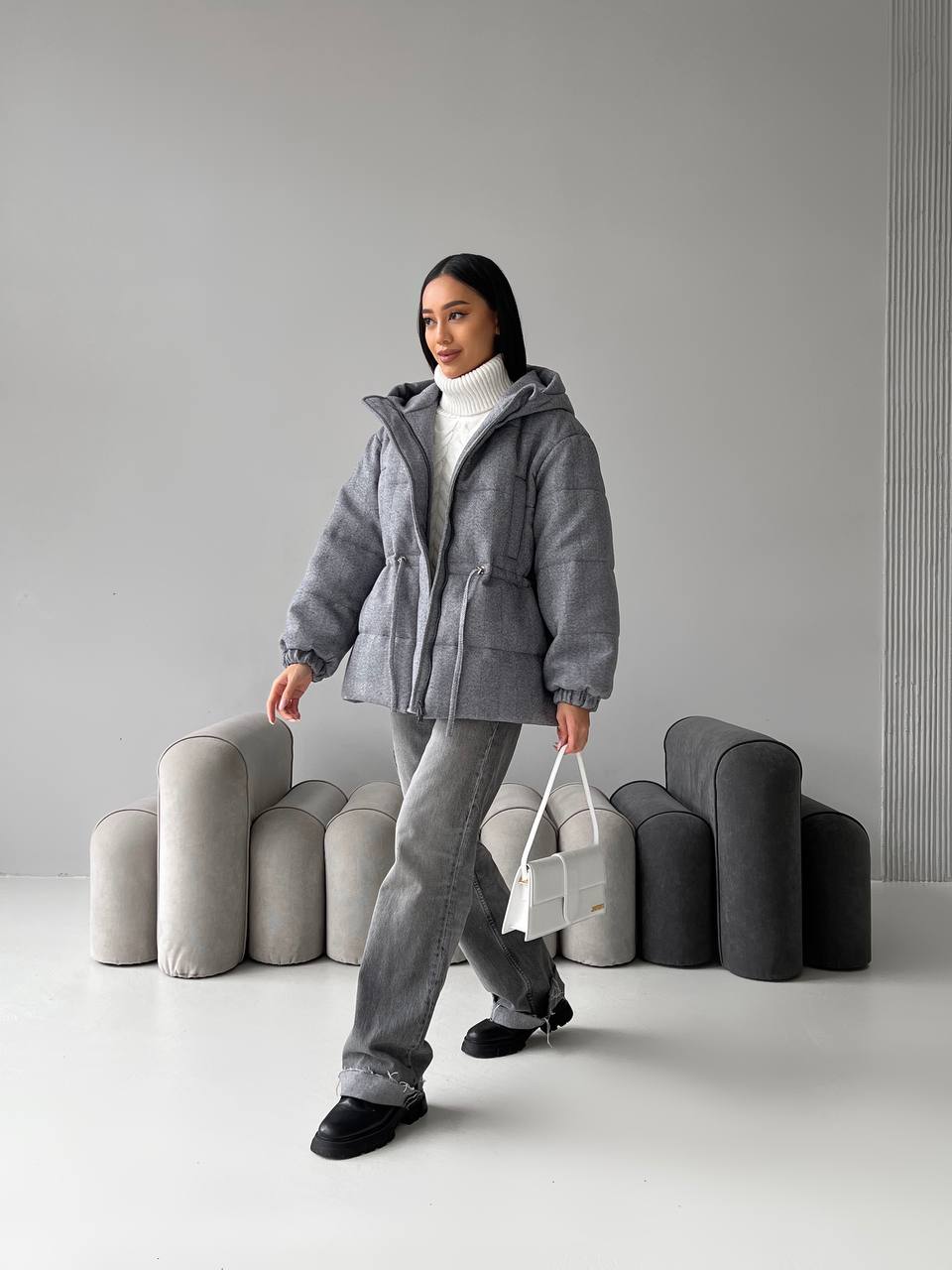 Person wearing a gray jacket and pants standing in a minimalistic room with gray furniture.