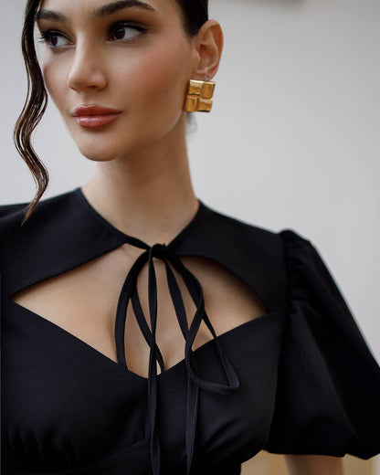 Woman wearing a black top with a tie detail and gold earrings against a neutral background