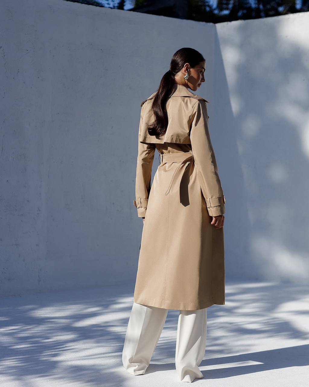 Woman wearing a beige trench coat against a white wall